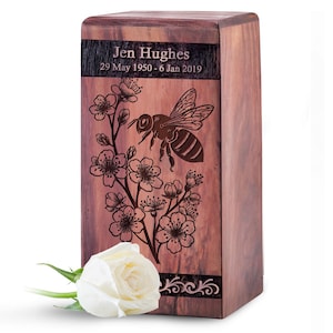 May include: A rectangular wooden cremation urn with a dark wood grain finish. The front features an engraved bee and cherry blossom branch design, with the text "Jen Hughes" and "29 May 1950 - 6 Jan 2019" at the top. A white rose rests at the base.