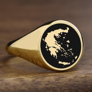 Peut inclure: Une bague chevalière dorée avec une face circulaire noire représentant une carte détaillée de la Grèce. La bague est posée sur une surface en bois, mettant en valeur sa finition polie et son design.