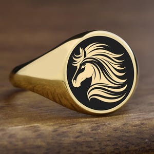 Op de afbeelding: Een goudkleurige zegelring met een zwarte, cirkelvormige voorkant met een goudkleurig paardenhoofdsilhouet. De ring ligt op een houten oppervlak, wat de gepolijste afwerking en het ontwerp benadrukt.
