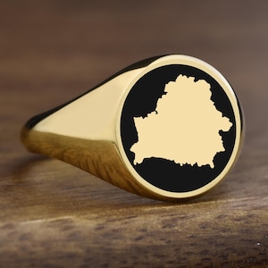 May include: A gold-colored signet ring with a black circular face. The face features a gold-colored outline of the country of Belarus. The ring is displayed on a wooden surface.