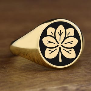 Op de afbeelding: Een gouden zegelring met een zwart cirkelvormig embleem met een gestileerd bladontwerp. De ring heeft een gepolijste afwerking en ligt op een houten oppervlak.