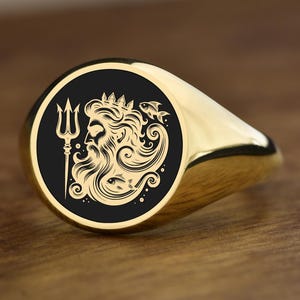 Pode incluir: Um anel de sinete dourado com uma face preta com uma gravura detalhada de uma figura barbuda com um tridente e uma coroa. O anel tem um acabamento polido e uma forma arredondada, sobre uma superfície de madeira.