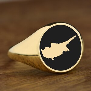 Op de afbeelding: Een gouden zegelring met een zwarte cirkelvormige face met een gouden omtrek van Cyprus. De ring ligt op een houten oppervlak.