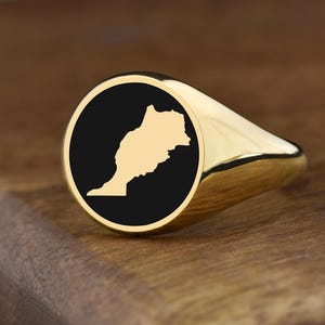 Può includere: Un anello con sigillo dorato con una faccia circolare nera che mostra un contorno dorato della mappa del Marocco. L'anello è presentato su una superficie di legno, evidenziando la sua finitura lucida e il suo design.