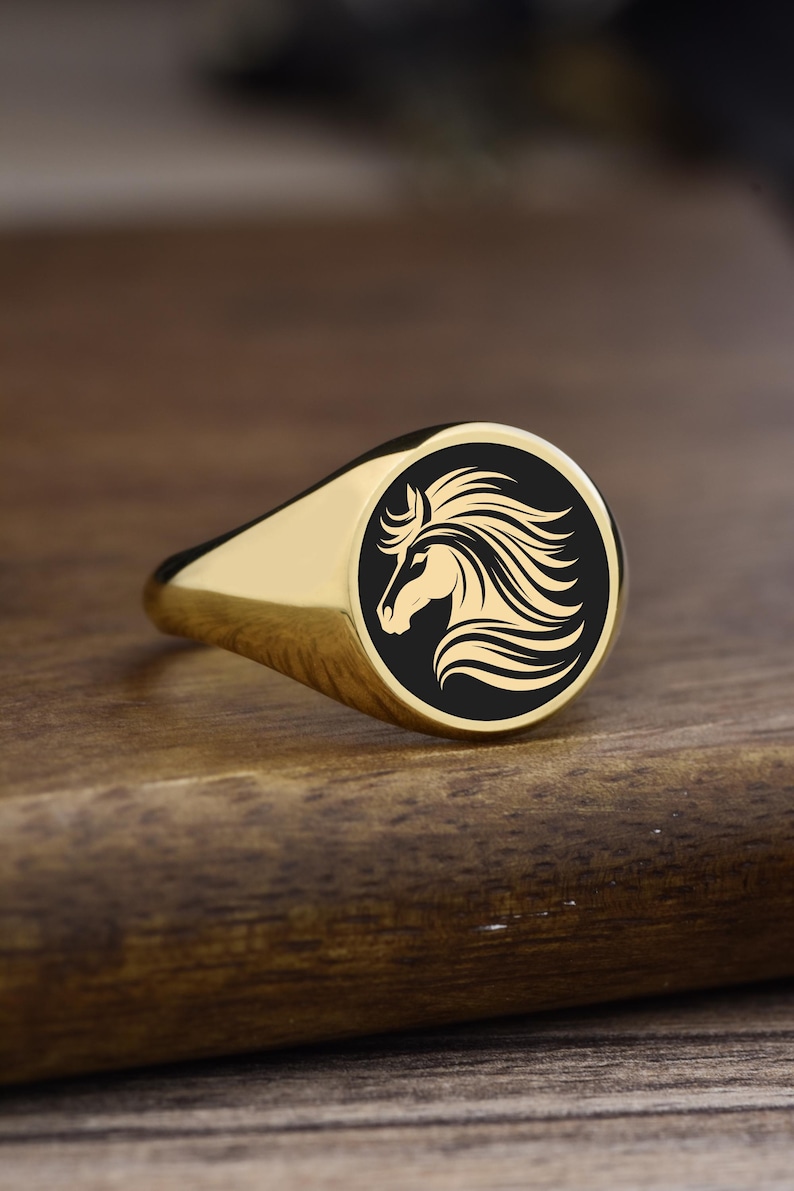 Op de afbeelding: Een goudkleurige zegelring met een zwarte, cirkelvormige voorkant met een goudkleurig paardenhoofdsilhouet. De ring ligt op een houten oppervlak, wat de gepolijste afwerking en het ontwerp benadrukt.
