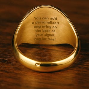 Op de afbeelding: Close-up van een gouden zegelring. De ring heeft een glad, gepolijst oppervlak en een afgeronde, ovale vorm. Op de achterkant van de ring staat de tekst gegraveerd: "You can add a personalized engraving on the back of your signet ring for free!"