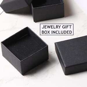 Op de afbeelding: Verschillende kleine, open zwarte sieraden geschenkdozen zijn gerangschikt op een lichtgekleurd oppervlak. De dozen zijn vierkant van vorm met een matte afwerking. De tekst "JEWELRY GIFT BOX INCLUDED" wordt weergegeven in een witte doos.