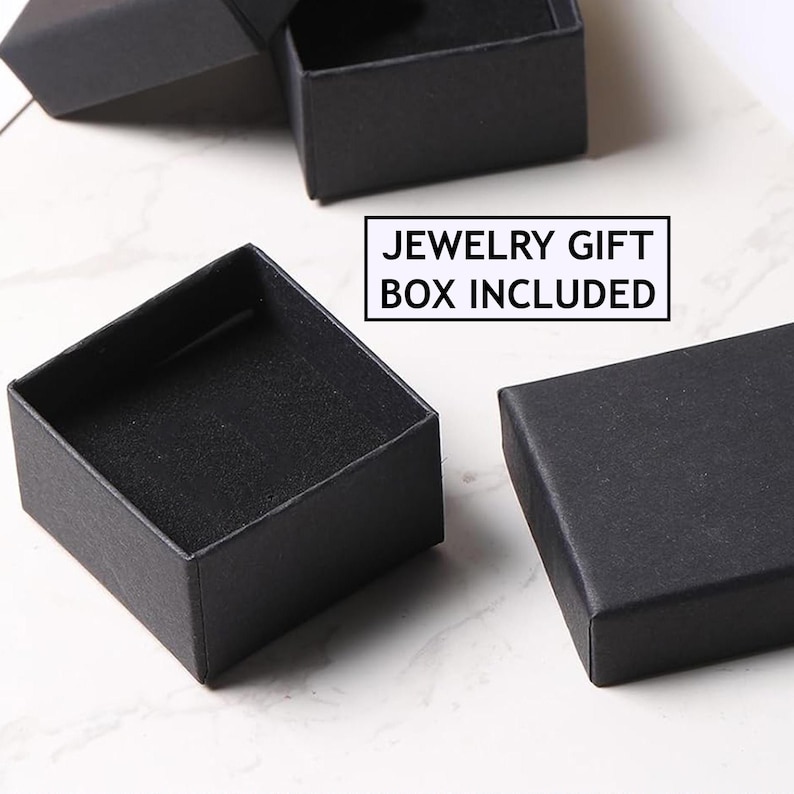 Op de afbeelding: Verschillende kleine, open zwarte sieraden geschenkdozen zijn gerangschikt op een lichtgekleurd oppervlak. De dozen zijn vierkant van vorm met een matte afwerking. De tekst "JEWELRY GIFT BOX INCLUDED" wordt weergegeven in een witte doos.