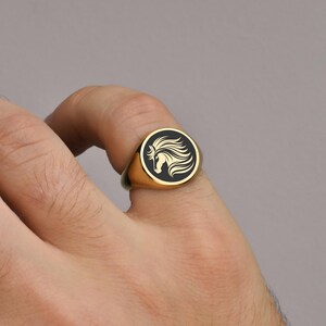 Op de afbeelding: Goudkleurige zegelring met een zwarte cirkelvormige face met een wit paardenkopontwerp. De ring wordt op een vinger getoond tegen een neutrale achtergrond. Een klassiek sieraad.