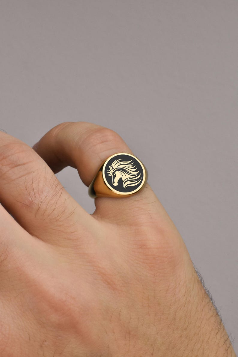 Op de afbeelding: Goudkleurige zegelring met een zwarte cirkelvormige face met een wit paardenkopontwerp. De ring wordt op een vinger getoond tegen een neutrale achtergrond. Een klassiek sieraad.