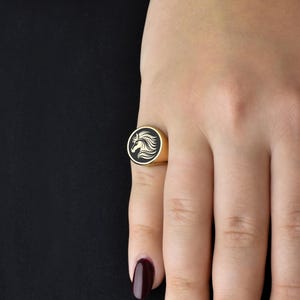 Op de afbeelding: Goudkleurige ring met een zwarte, cirkelvormige voorkant met een wit paardenhoofd ontwerp. De ring is rond en zit om een vinger. De achtergrond is zwart.
