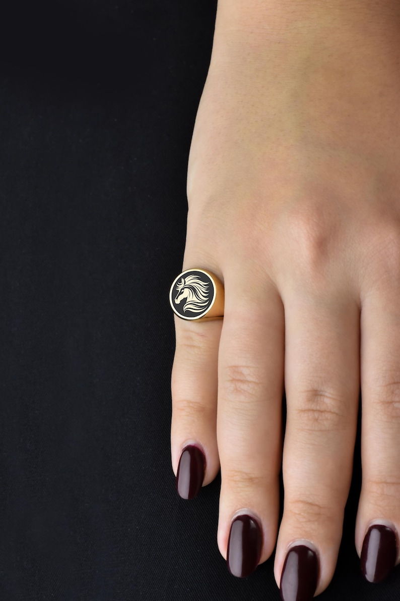 Op de afbeelding: Goudkleurige ring met een zwarte, cirkelvormige voorkant met een wit paardenhoofd ontwerp. De ring is rond en zit om een vinger. De achtergrond is zwart.