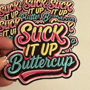 May include: A fabric patch featuring embroidered text that reads SUCK IT UP BUTTERCUP. The word SUCK is in pink and yellow, IT UP is in yellow, and BUTTERCUP is in light blue. The entire design is outlined in black with a pink border.