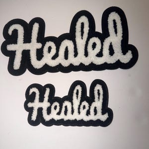 May include: Two black and white patches with the word "Healed" in a script font. The word is in white, with a black border. One patch is larger than the other.