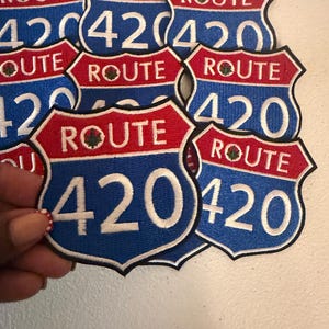 May include: A collection of shield-shaped embroidered patches featuring a blue background with the white number 420 and a red top section with the white text ROUTE and a small green leaf icon. The patches have a black border and white stitching.