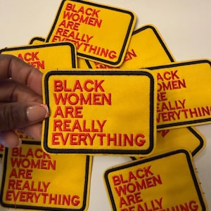 May include: Yellow rectangular patches with black borders and the red text "BLACK WOMEN ARE REALLY EVERYTHING." Several patches are visible, with one held by a hand. The patches are embroidered.