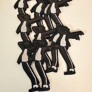 May include: Black and white embroidered patches in the shape of a person in a suit, with arms outstretched. The patches feature a black suit with white accents and a white shirt. The patches are stacked on top of each other.