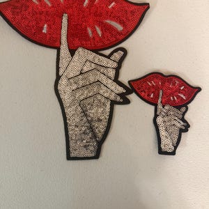 May include: Two sequined art pieces: a large red sequined mouth with a finger to the lips, and a smaller version. The hands are silver sequins with black outlines. The background is a light gray.