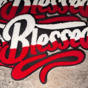 Puede incluir: Primer plano de varios parches bordados con la palabra "Blessed" en una fuente de escritura. Los parches presentan un fondo negro con letras blancas, rojas y negras. Los parches son probablemente para ropa o accesorios.