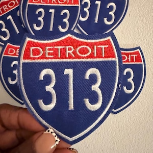 May include: A shield-shaped embroidered patch featuring a blue background with a red horizontal band at the top. The word DETROIT is stitched in white within the red band, and the number 313 is stitched in large white numerals below.