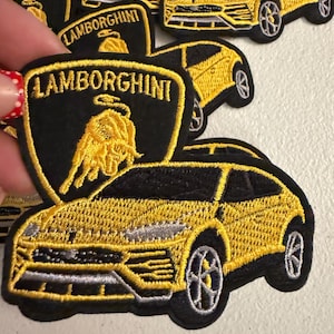 May include: A collection of embroidered patches featuring a yellow Lamborghini car and the black and yellow Lamborghini shield logo with the text LAMBORGHINI. The patches are stitched with yellow, black, and silver thread on a black fabric base.