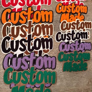 May include: A collection of custom-made embroidered patches in various colors. The patches feature the word "Custom" in red, orange, brown, black, purple, and green, with some including "Made". The patches are on a wooden surface.