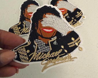 Puede incluir: Tres parches bordados con un retrato estilizado de Michael Jackson. El diseño muestra una figura con cabello oscuro, una sonrisa roja y un guante de lentejuelas plateadas cubriendo los ojos. La chaqueta es negra con detalles dorados y el nombre Michael Jackson.