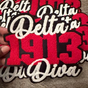 May include: Red and black embroidered patches with the words "Delta 1913" in white script. The patches have a textured, felt-like appearance. The word "Diva" is also visible on one of the patches.