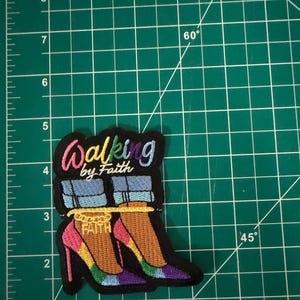 May include: An embroidered patch featuring the text Walking by Faith in colorful lettering above an illustration of two feet wearing blue jeans and high-heeled shoes with a gold anklet that reads FAITH, measuring approximately 3.5 inches tall.