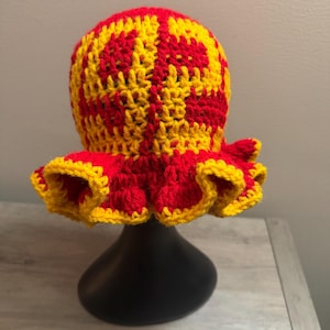 May include: A handmade crochet hat in red and yellow. The hat features a checkerboard pattern on the crown and a ruffled brim. The hat is displayed on a black stand.