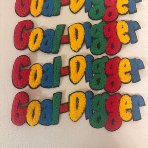 May include: Four colorful embroidered patches with the text "Goal Digger". Each patch features the words in red, yellow, green, and blue, with a black border. The patches are arranged in a vertical stack.