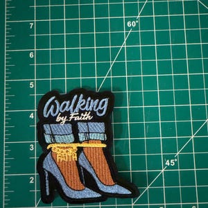 May include: An embroidered patch featuring light blue high-heeled shoes with cuffed denim jeans, a gold chain anklet with the word FAITH, and the text Walking by Faith in light blue script, measuring approximately 3.5 inches in height.
