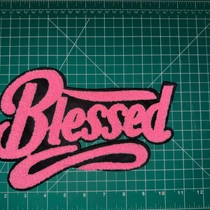 Puede incluir: Un parche bordado rosa y negro con la palabra "Blessed" en una fuente cursiva. El parche tiene una textura difusa y est&aacute; colocado sobre una esterilla de corte verde.