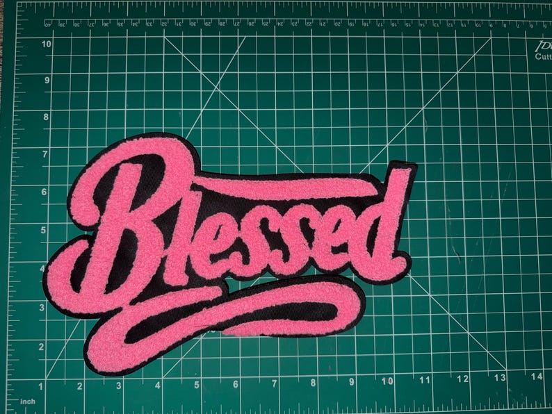 Puede incluir: Un parche bordado rosa y negro con la palabra "Blessed" en una fuente cursiva. El parche tiene una textura difusa y est&aacute; colocado sobre una esterilla de corte verde.