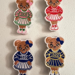 May include: Four embroidered bear patches featuring brown bears wearing sweaters and pleated skirts. The top left bear wears pink with 1908, top right wears red with 1913, bottom left wears blue with 1920, and bottom right wears blue and gold with 1922.