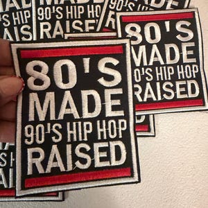 May include: Black square patches with white embroidered text that reads "80'S MADE 90'S HIP HOP RAISED". The patches have a red border and are stacked on top of each other.