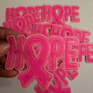 May include: A collection of bright pink embroidered patches featuring the word HOPE, where the letter O is replaced by a breast cancer awareness ribbon. Each patch has a textured, dotted surface and a stitched border, stacked together in a pile.