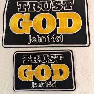 May include: Two black rectangular patches with white borders. The top patch reads "TRUST GOD" in white block letters above the yellow embroidered word "GOD". Below is "John 14:1". The bottom patch is a smaller version of the top patch.