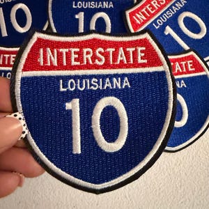 May include: A shield-shaped embroidered patch featuring the text INTERSTATE in white on a red background, with LOUISIANA and the number 10 in white on a blue background. The patch has a black border and is part of a collection of identical items.