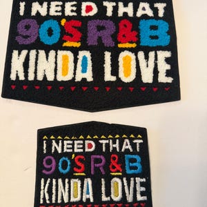 May include: Two black patches with the text "I NEED THAT 90'S R&B KINDA LOVE" in colorful, embroidered letters. The patches have a unique shape with a pointed bottom and a decorative border.