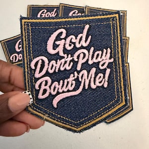 May include: A dark blue denim patch shaped like a pocket with gold stitching along the edges. The patch features the embroidered text "God Don't Play Bout' Me!" in a light pink cursive font. The design mimics the appearance of a jean pocket.