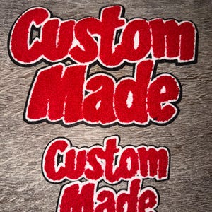 May include: Two red felt patches with the words "Custom Made" in a bold, cursive font. The text is outlined in white and black. The patches are displayed on a wooden surface.