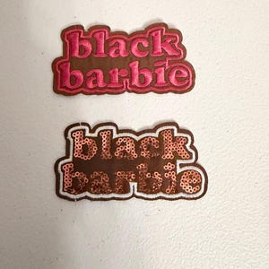 May include: Two embroidered patches with the text "black barbie". One patch has pink lettering on a brown background. The other patch has sequined lettering on a brown background with a white border.