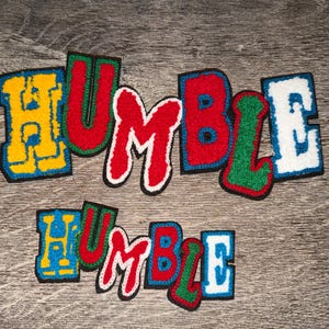 May include: Colorful embroidered patches spelling the word "HUMBLE". The patches feature a variety of colors, including yellow, red, green, blue, and white, with black borders. The patches are arranged on a wooden surface.
