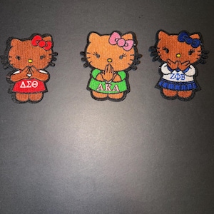 May include: Three embroidered patches featuring a cartoon cat with a bow. Each cat wears a different outfit and has a different Greek letter combination: Delta Sigma Theta, Alpha Kappa Alpha, and Zeta Phi Beta. The patches are brown, red, green, blue, and white.