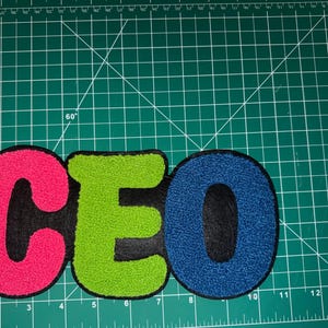 Peut inclure: Un &eacute;cusson color&eacute; avec les lettres "CEO" en rose vif, vert et bleu. Les lettres sont brod&eacute;es avec un aspect textur&eacute; et flou et sont soulign&eacute;es en noir. L'&eacute;cusson est sur un tapis de coupe vert.