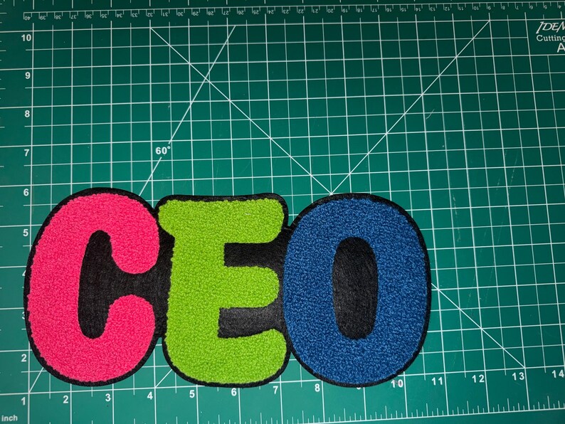 Peut inclure: Un &eacute;cusson color&eacute; avec les lettres "CEO" en rose vif, vert et bleu. Les lettres sont brod&eacute;es avec un aspect textur&eacute; et flou et sont soulign&eacute;es en noir. L'&eacute;cusson est sur un tapis de coupe vert.