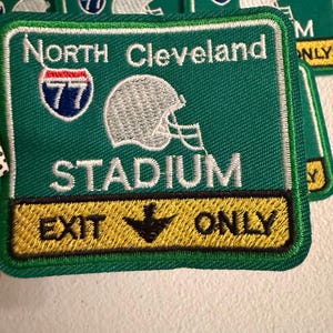 May include: A square embroidered patch with a green background featuring the text North Cleveland Stadium in white. It includes a white football helmet graphic, a blue and red interstate 77 shield, and a yellow bottom bar with black text Exit Only.