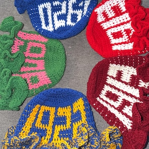May include: Five handmade crochet hats in various colors: blue, red, green, and burgundy. Each hat features a unique design with white or yellow lettering. The hats have a ruffled brim.