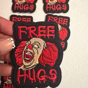 May include: A black embroidered patch featuring a clown face with red hair and a red nose, with the text "FREE HUGS" in red, dripping-style lettering above and below the clown face. The patch has a black border and a textured fabric base.